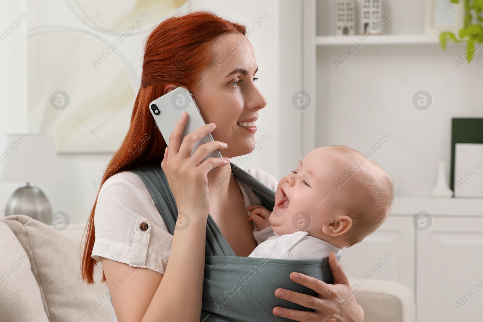 Photo of Mother talking on smartphone while holding her child in sling (baby carrier) at home