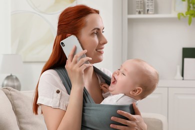 Mother talking on smartphone while holding her child in sling (baby carrier) at home