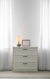 Modern chest of drawers with lamp near window in room