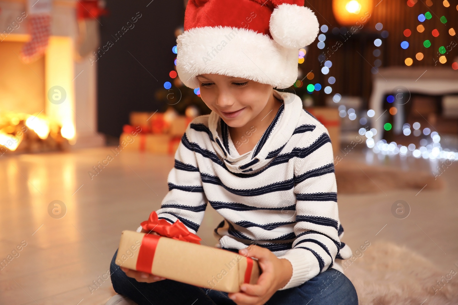 Photo of Cute little child in Santa hat with Christmas gift box at home