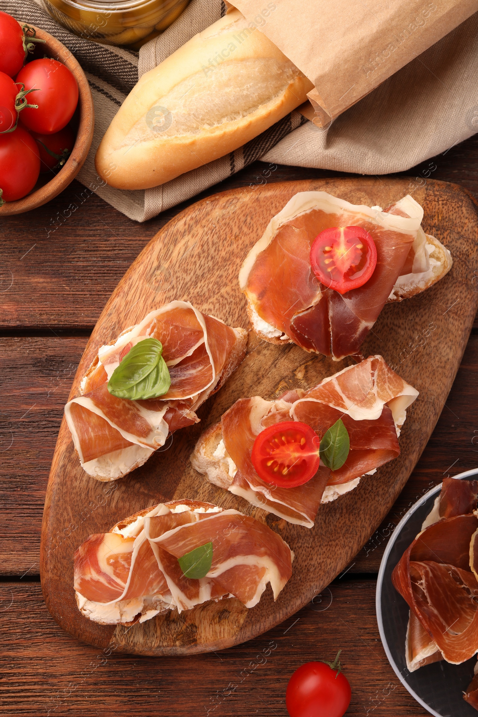 Photo of Board of tasty sandwiches with cured ham, basil and tomatoes on wooden table, flat lay