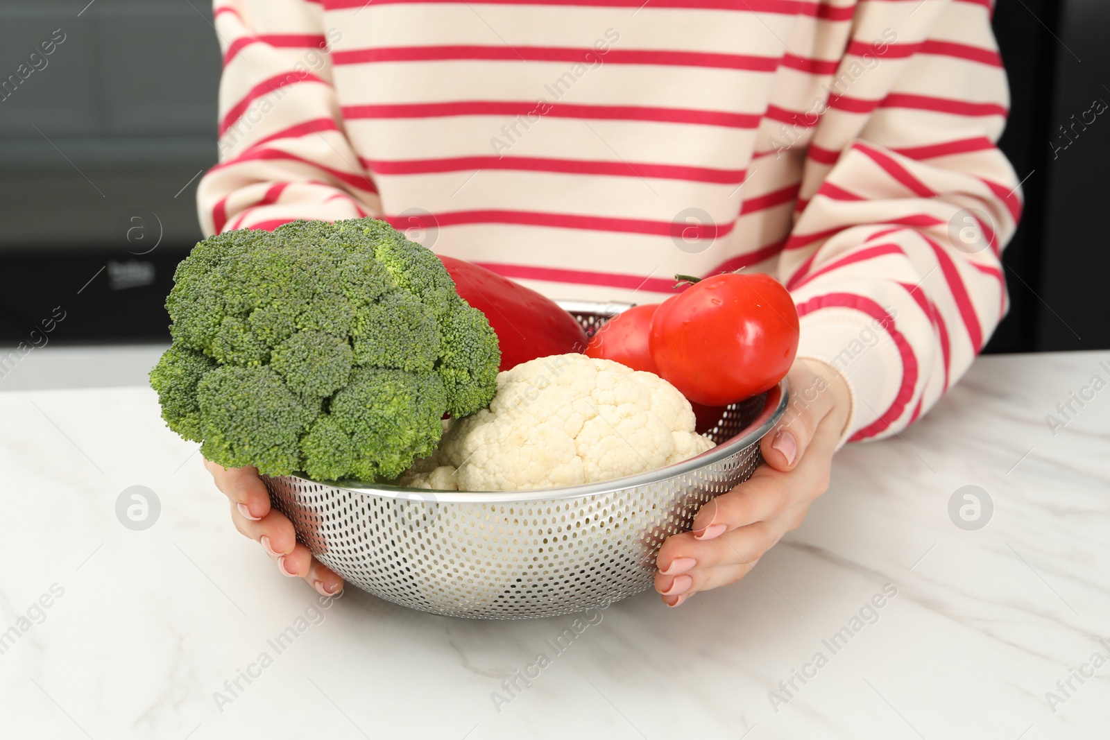 Photo of Woman holding colander with fresh vegetables at white marble table in kitchen, closeup