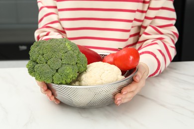 Woman holding colander with fresh vegetables at white marble table in kitchen, closeup