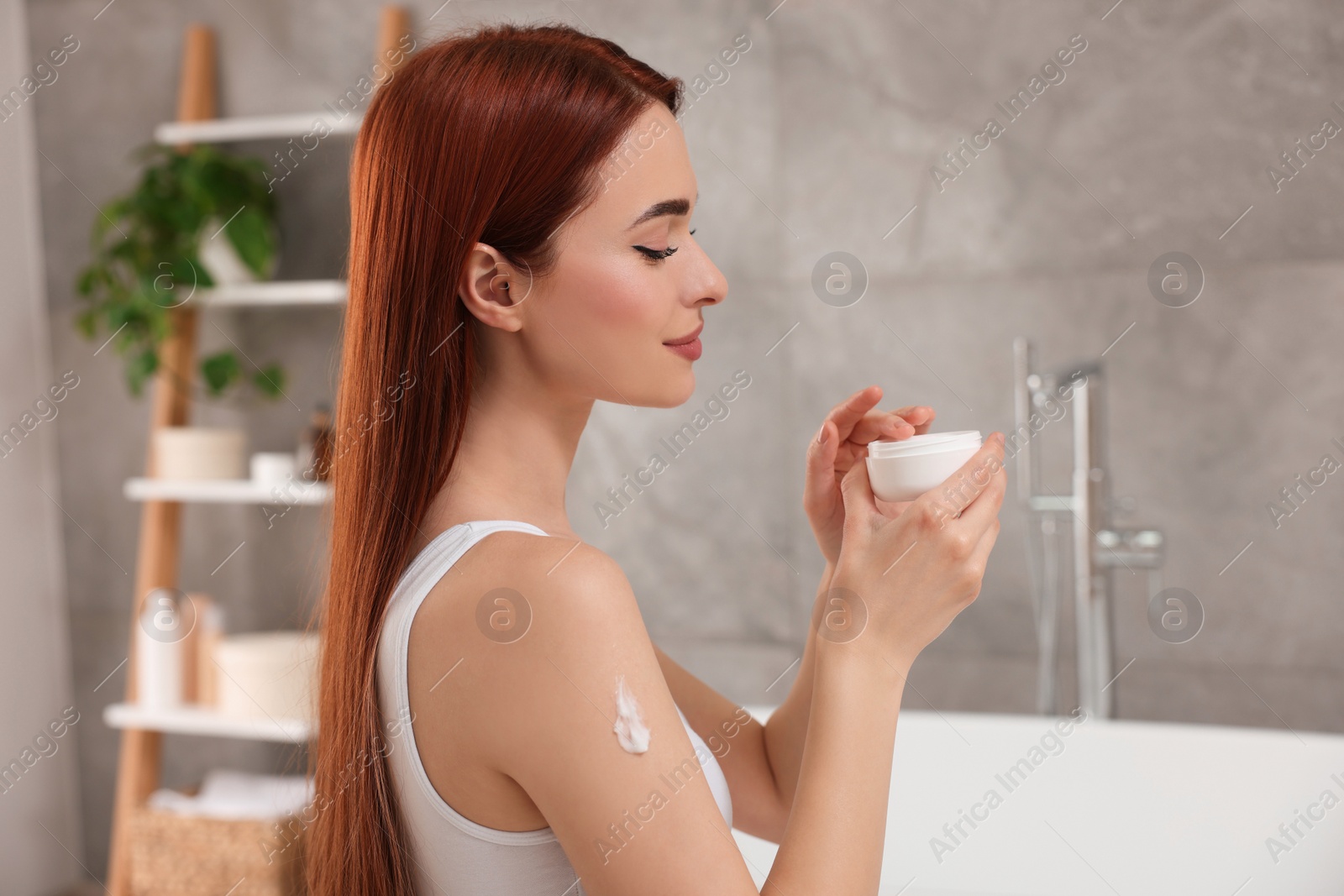 Photo of Beautiful young woman applying body cream onto shoulder in bathroom