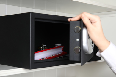 Photo of Woman opening black steel safe with money and jewelry on shelf, closeup
