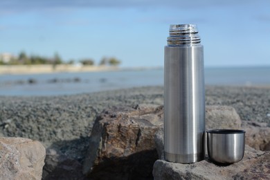 Photo of Metallic thermos and cap on stone near sea, space for text