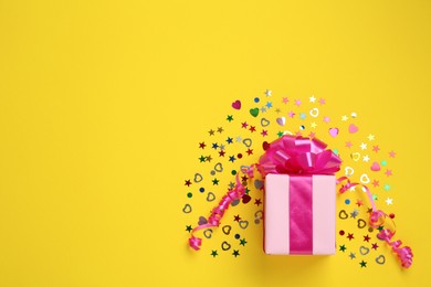 Photo of Pink gift box and shiny confetti on yellow background, top view. Space for text