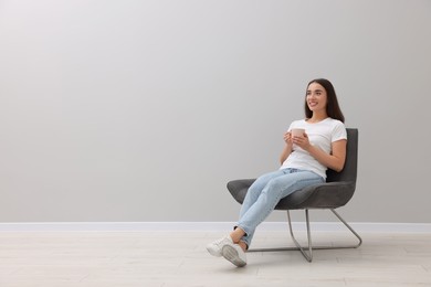 Beautiful woman with cup of drink sitting in armchair near light grey wall indoors, space for text