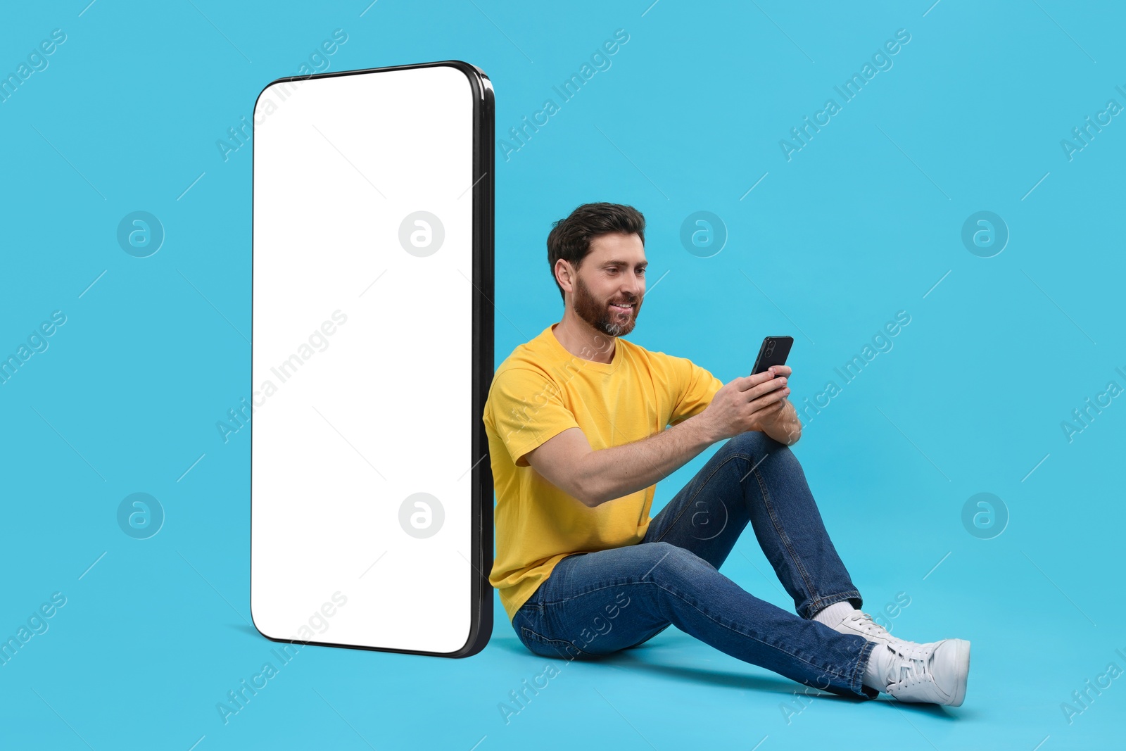 Image of Man with mobile phone sitting near huge device with empty screen on light blue background. Mockup for design