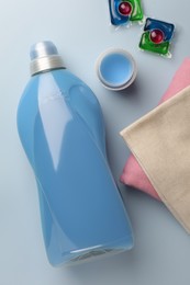 Bottle of fabric softener, laundry capsules and stacked clean clothes on light background, flat lay