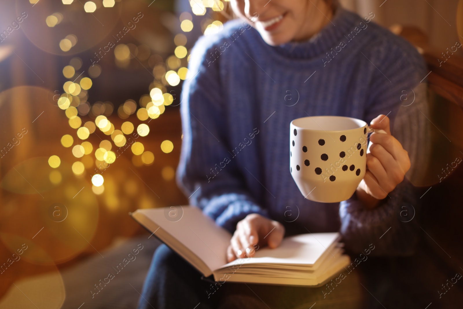 Photo of Woman with cup of hot beverage reading book at home in winter evening, closeup