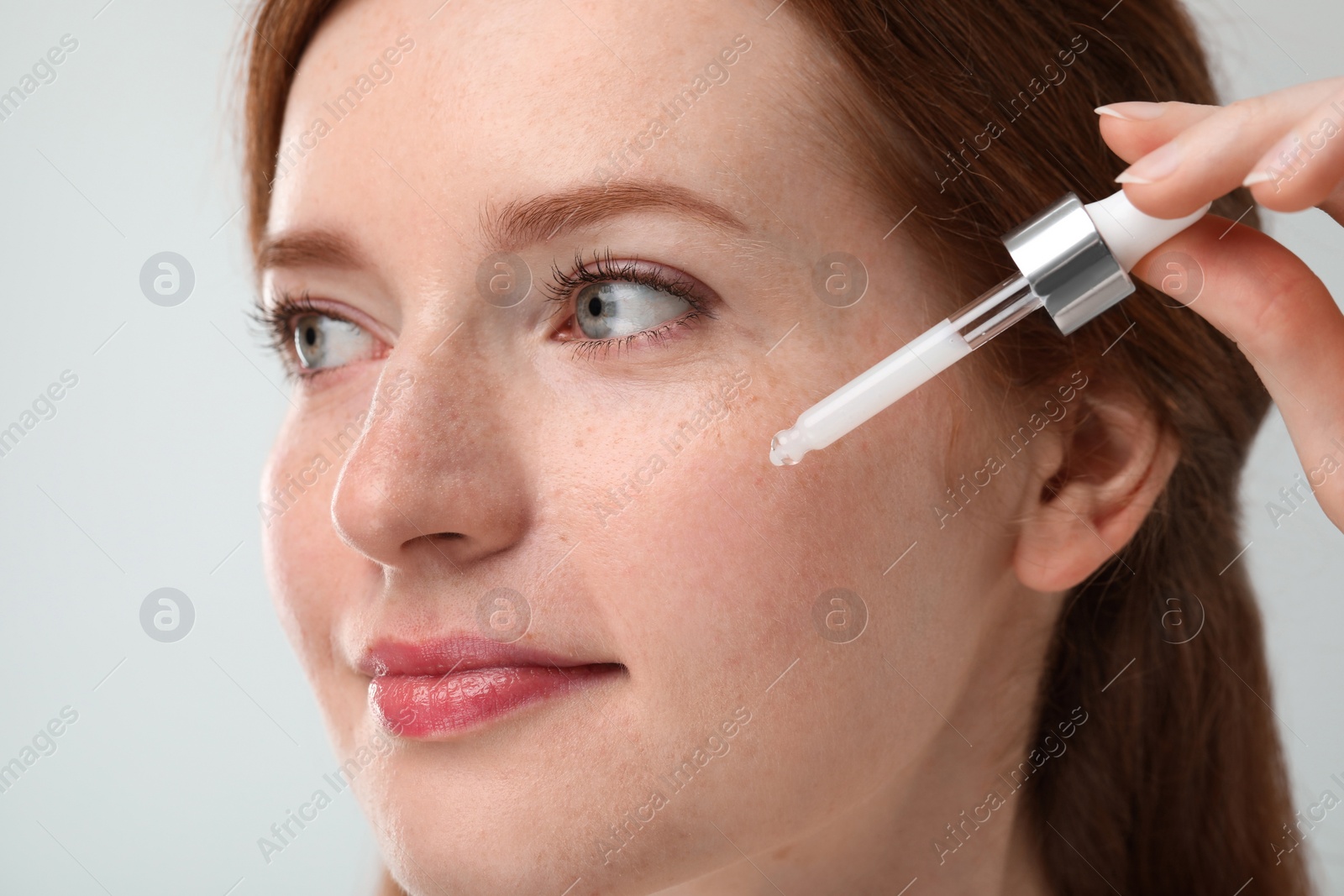 Photo of Beautiful woman with freckles applying cosmetic serum onto her face against grey background, closeup