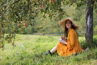 Photo of Beautiful young woman drawing with pencil in notepad on green grass near apple tree