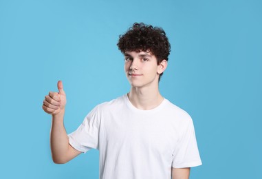 Portrait of cute teenage boy showing thumb up on light blue background