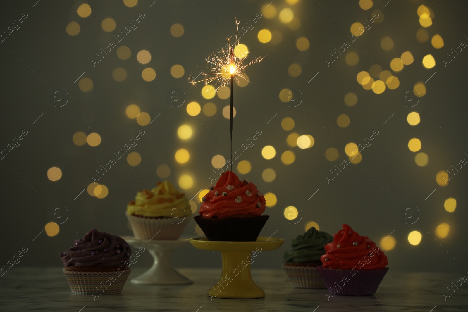 Photo of Different colorful cupcakes and one with sparkler on table against blurred lights