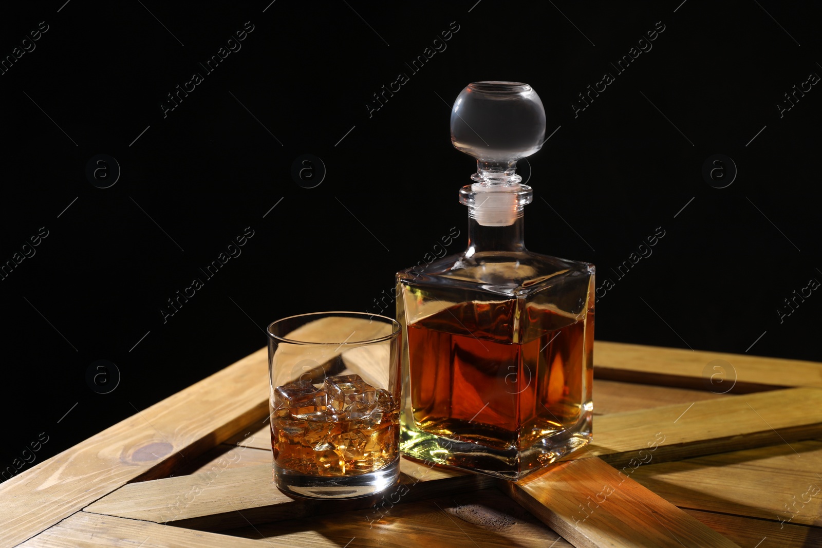 Photo of Whiskey with ice cubes in glass and bottle on wooden crate against black background