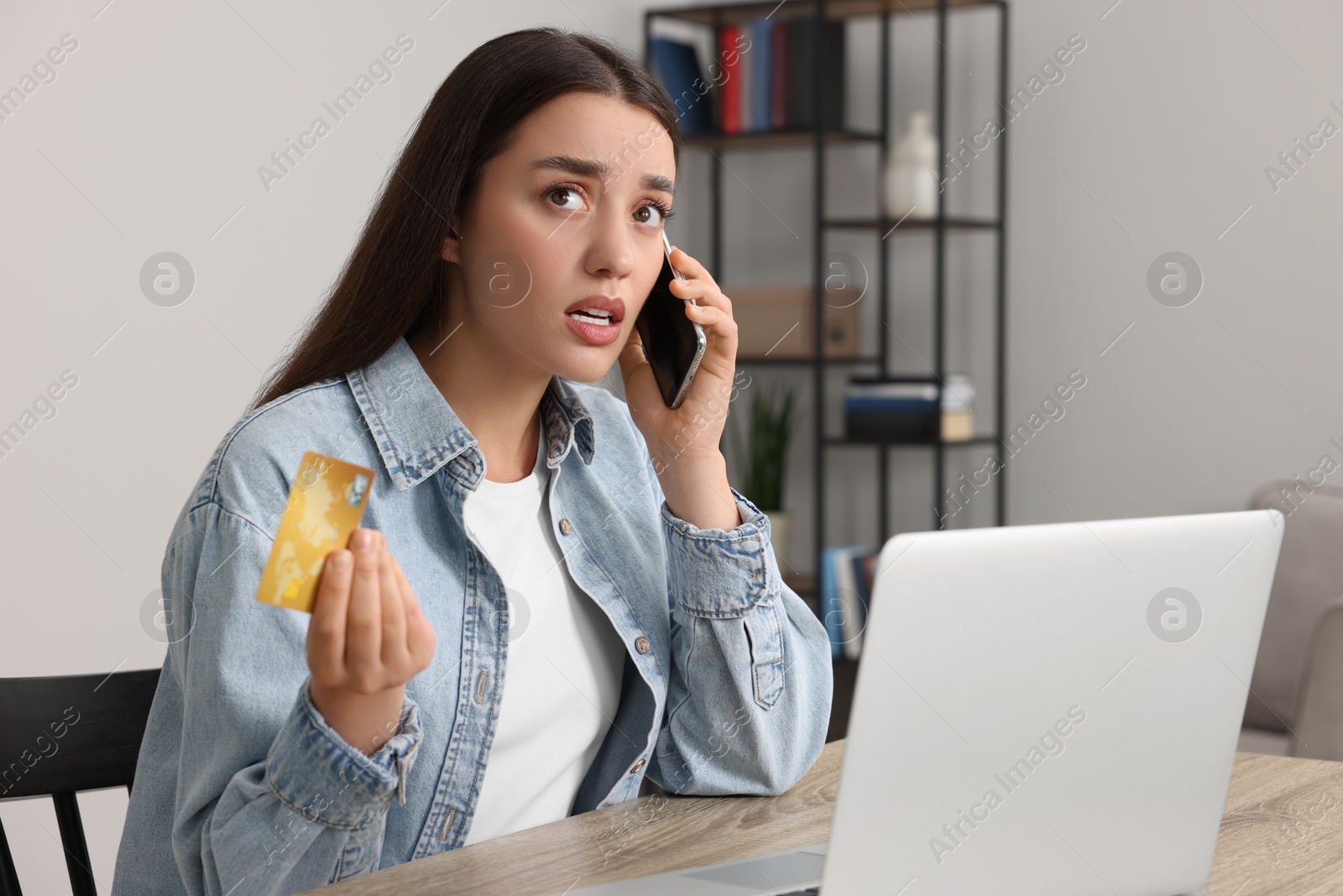 Photo of Confused woman with credit card talking on phone and laptop at table indoors. Be careful - fraud