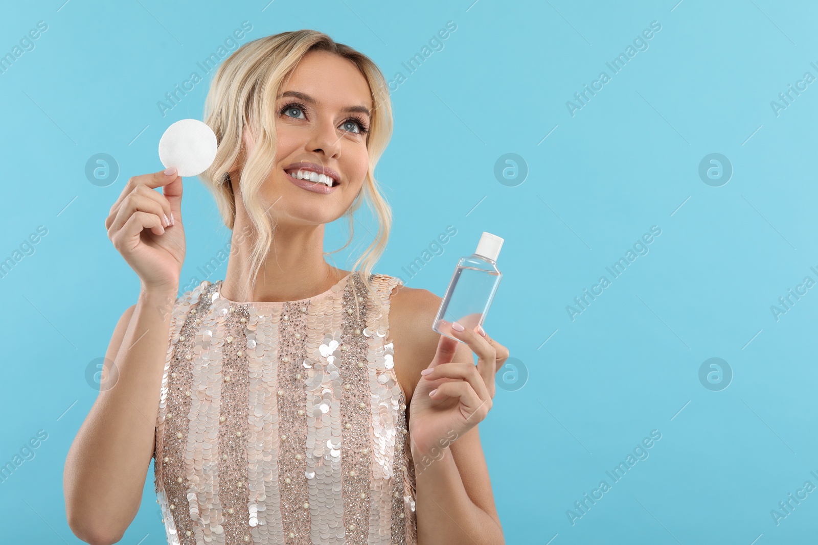 Photo of Smiling woman removing makeup with cotton pad and holding bottle on light blue background. Space for text
