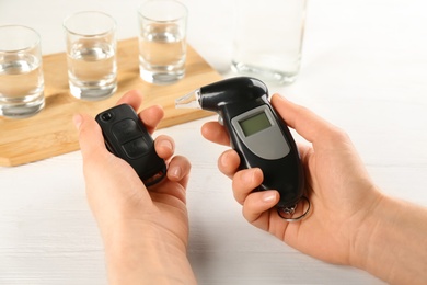 Woman holding modern breathalyzer and car key near white table with alcohol, closeup