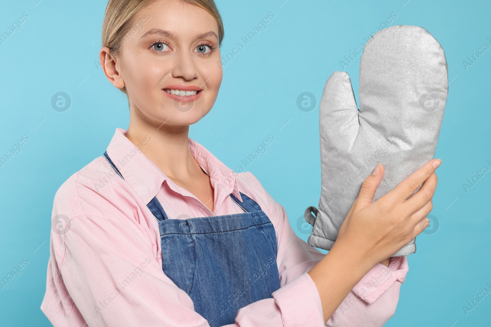 Photo of Beautiful young woman in denim apron and oven glove on light blue background, closeup