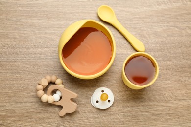 Photo of Pacifier, toy and plastic dishware with healthy baby food on wooden table, flat lay