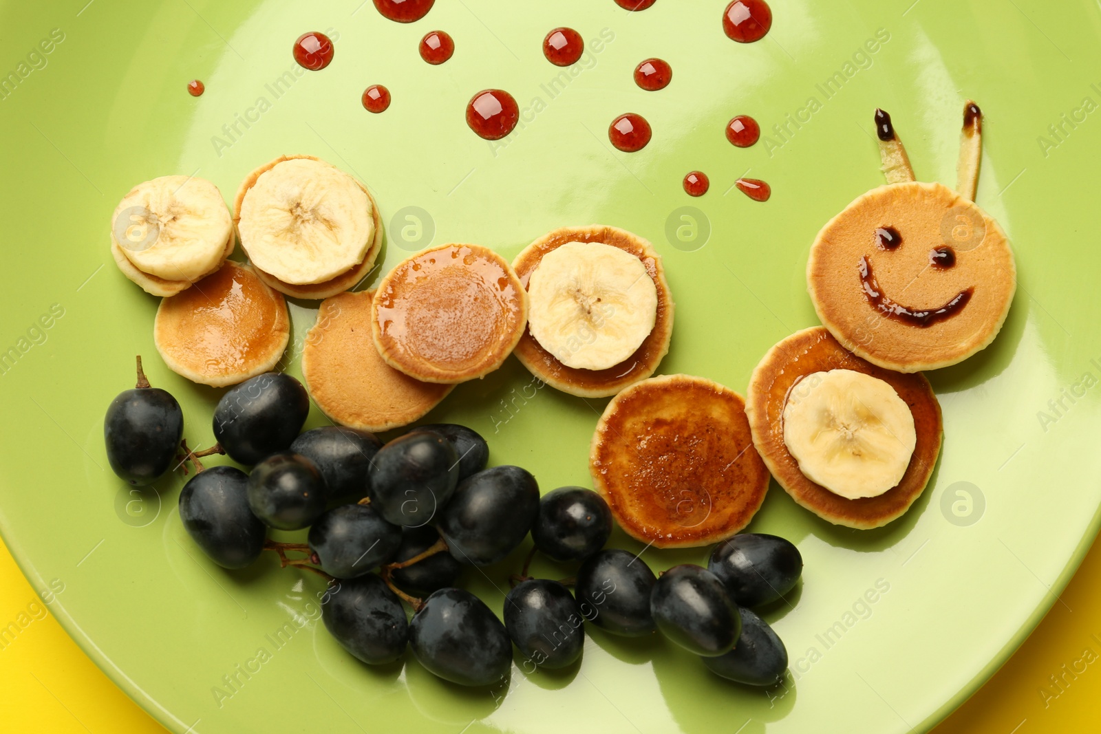 Photo of Creative serving for kids. Plate with cute caterpillar made of pancakes, grapes and banana on yellow background, top view