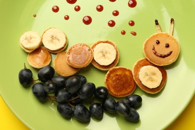 Photo of Creative serving for kids. Plate with cute caterpillar made of pancakes, grapes and banana on yellow background, top view