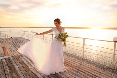 Photo of Gorgeous bride in beautiful wedding dress with bouquet near river on sunset