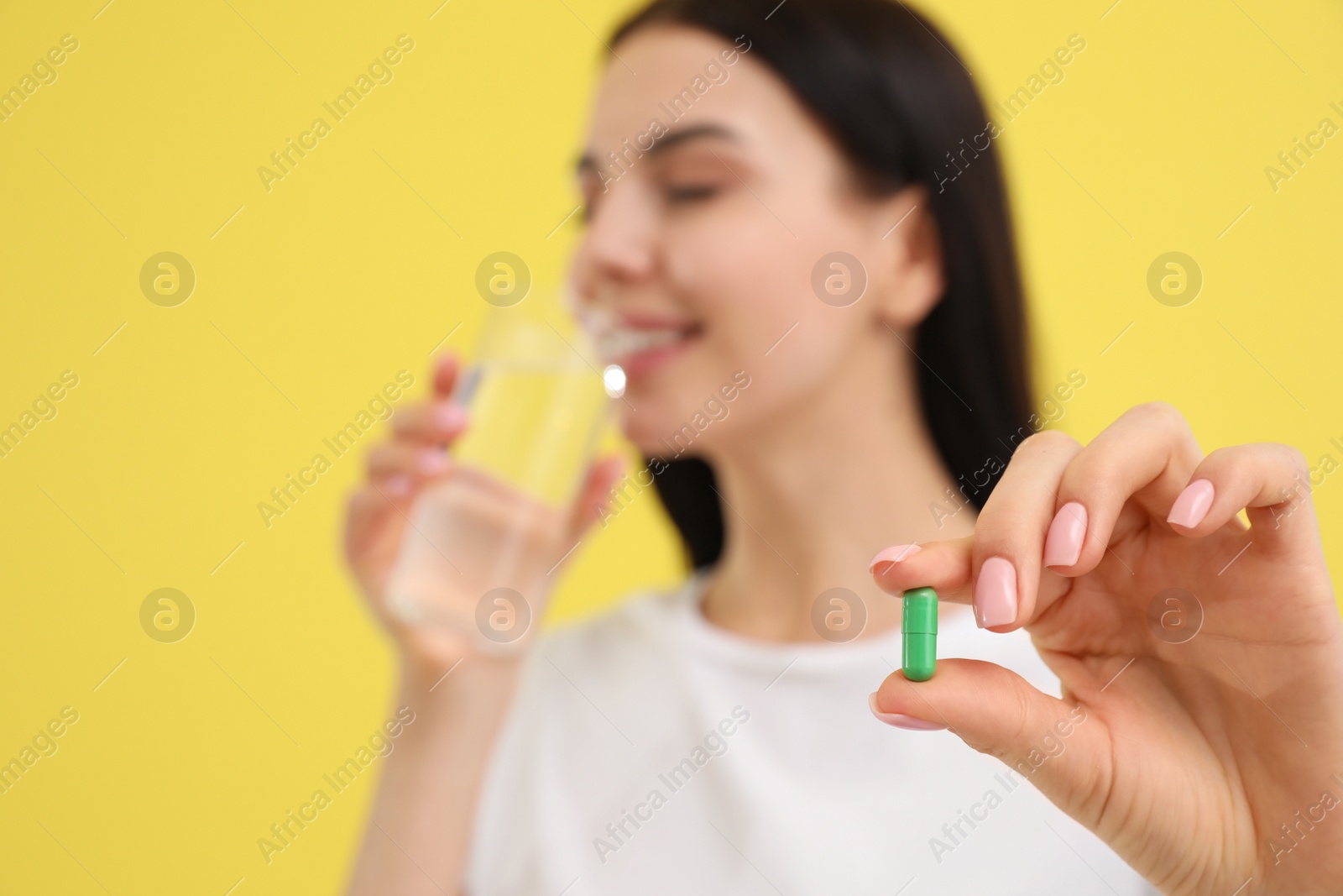 Photo of Young woman with glass of water and vitamin capsule against yellow background, focus on hand