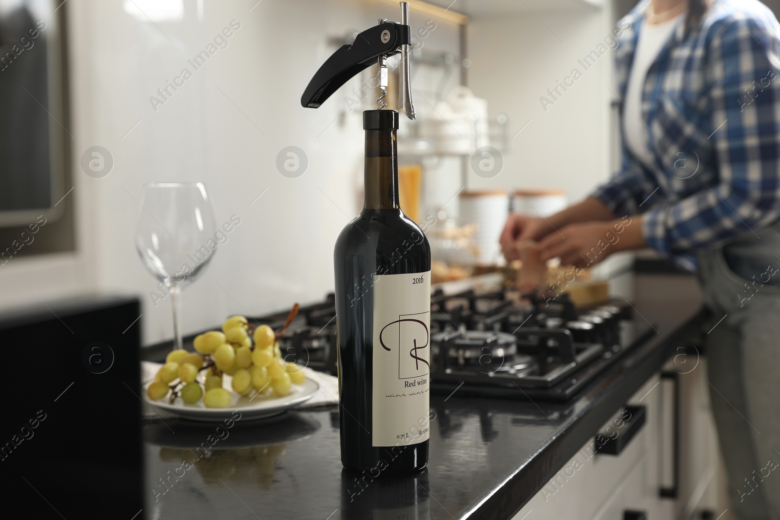 Photo of Wine bottle with corkscrew on black countertop, closeup. Woman in kitchen, selective focus