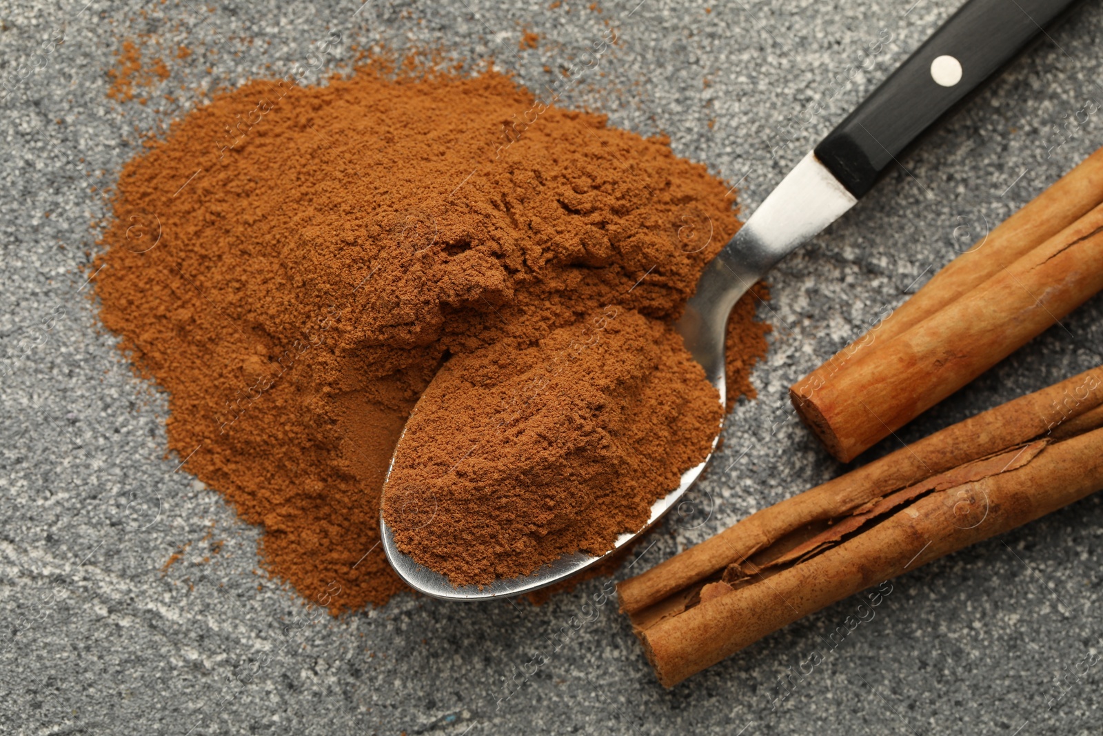 Photo of Spoon with cinnamon powder and sticks on grey table, flat lay