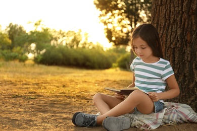 Photo of Cute little girl reading book near tree in park