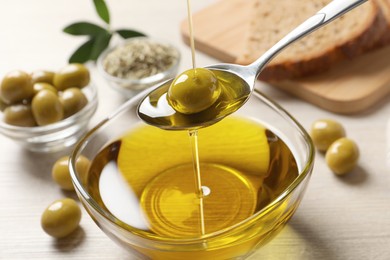 Photo of Pouring fresh olive oil into spoon above bowl on table, closeup