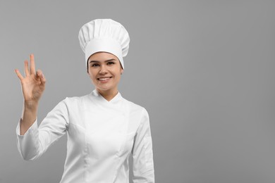 Photo of Happy female chef wearing uniform and cap showing ok gesture on light grey background. Space for text