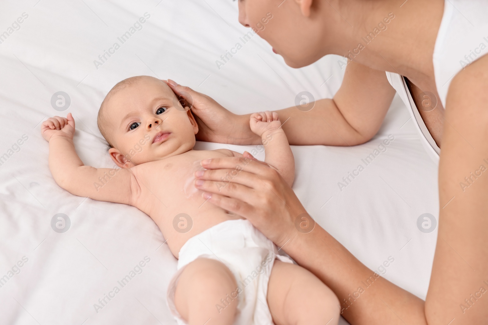 Photo of Woman applying body cream onto baby`s skin on bed, closeup