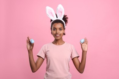 Photo of Happy African American woman in bunny ears headband holding Easter eggs on pink background