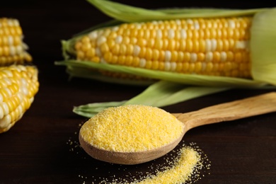 Photo of Cornmeal in spoon and fresh cobs on wooden table, closeup