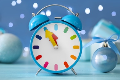Alarm clock with decor on light blue wooden table against blurred Christmas lights, closeup. New Year countdown