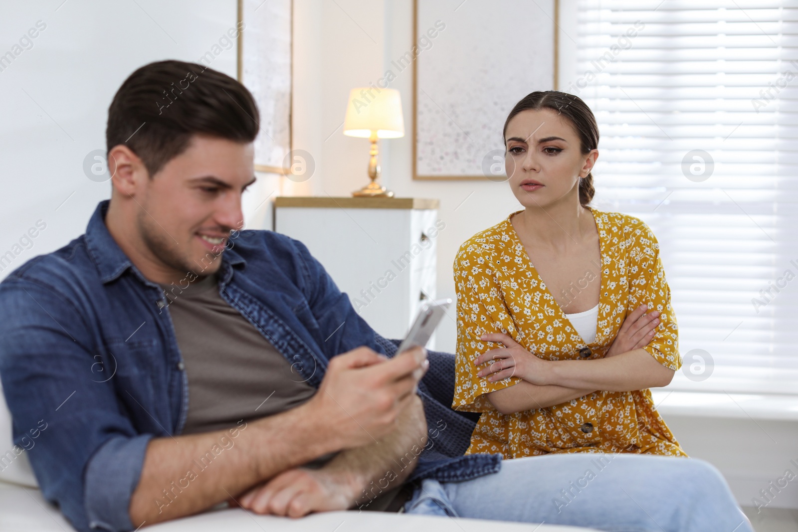 Photo of Distrustful woman peering into boyfriend's smartphone at home. Jealousy in relationship