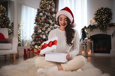 Photo of Young woman wearing Santa hat opening Christmas gift on floor at home