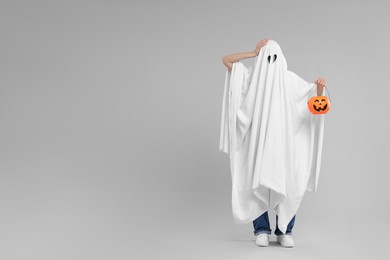 Photo of Woman in white ghost costume holding pumpkin bucket on light grey background, space for text. Halloween celebration