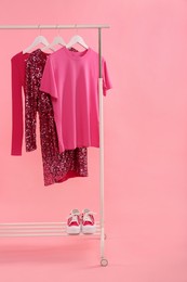 Rack with different stylish women`s clothes and sneakers on pink background, space for text
