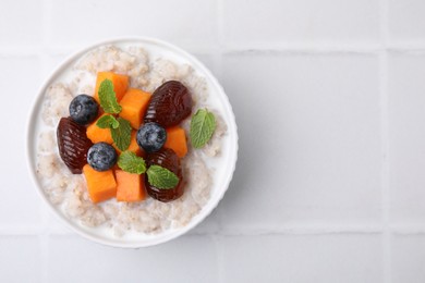 Delicious barley porridge with blueberries, pumpkin, dates and mint in bowl on white tiled table, top view. Space for text