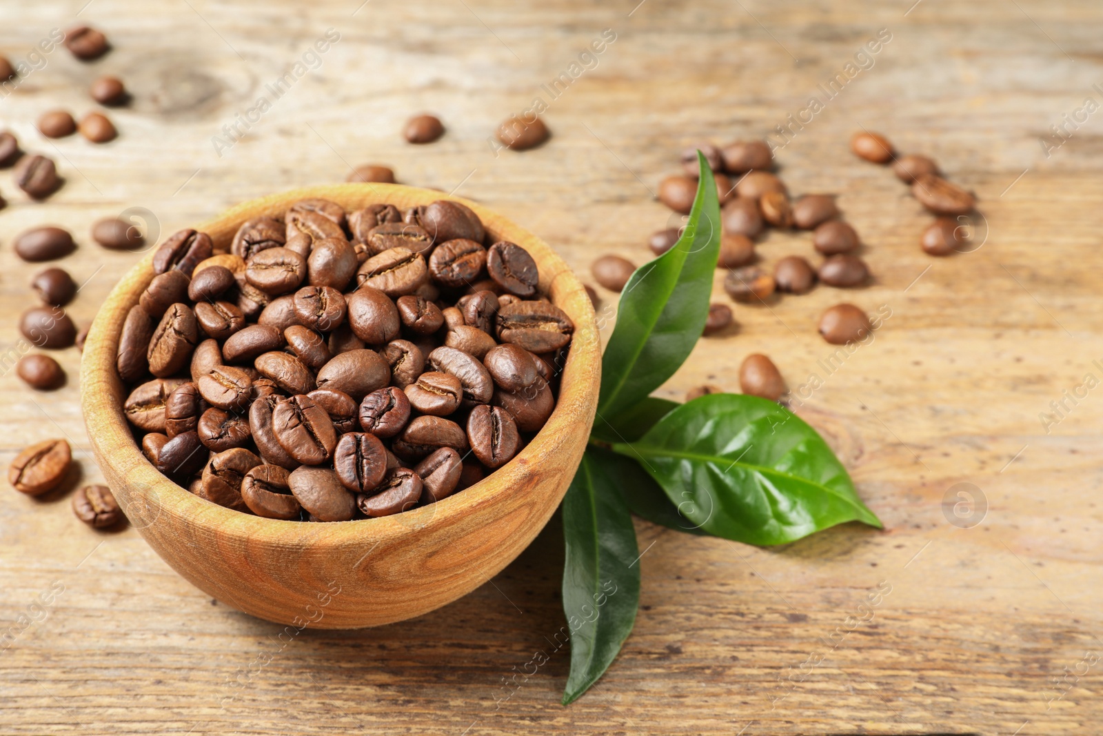 Photo of Bowl of coffee beans and fresh green leaves on wooden table, space for text