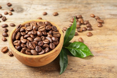 Photo of Bowl of coffee beans and fresh green leaves on wooden table, space for text