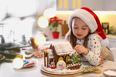 Photo of Cute little girl decorating gingerbread house at table indoors