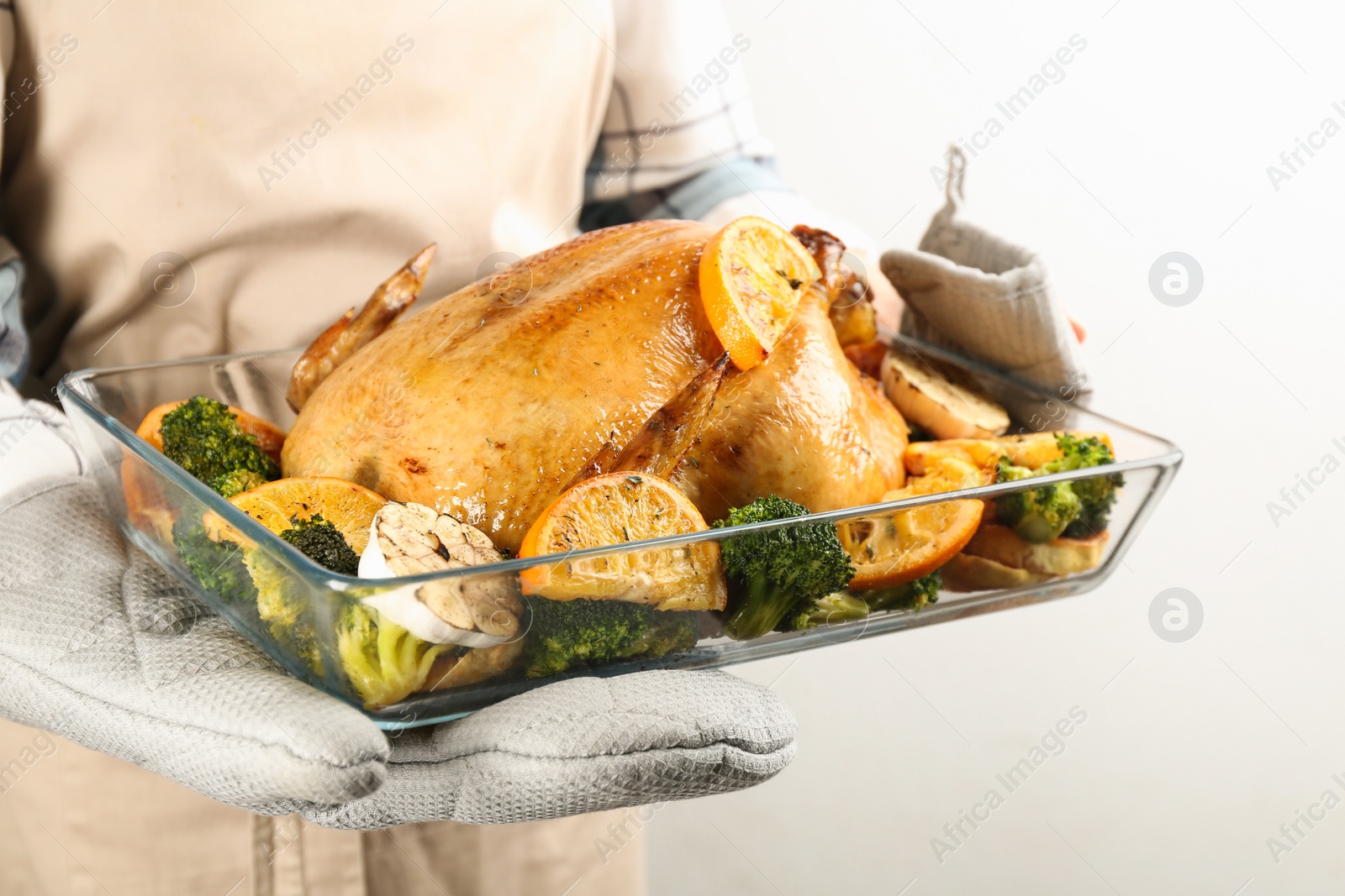 Photo of Woman holding pan with chicken, oranges and vegetables on light background, closeup