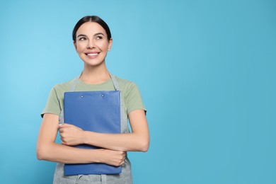 Young woman in grey apron with clipboard on light blue background, space for text