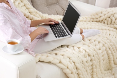 Photo of Woman using laptop on couch with soft knitted blanket at home, closeup