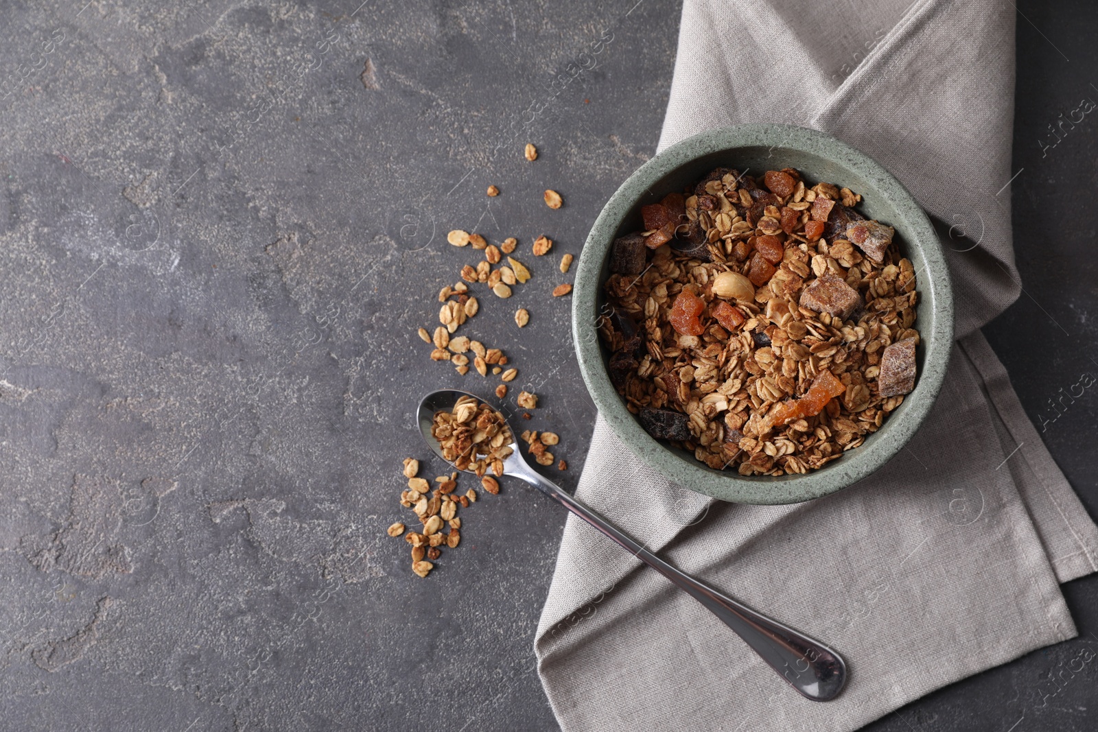 Photo of Tasty granola in bowl, spoon and napkin on gray textured table, flat lay. Space for text